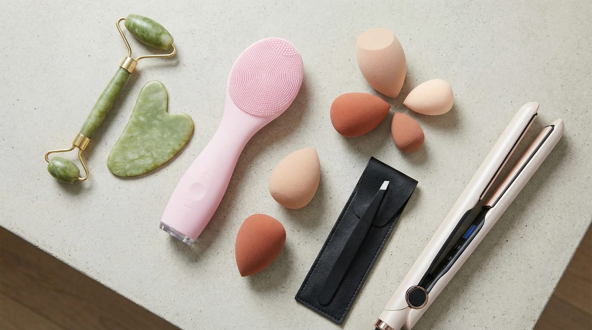 The Best Beauty Tools Every Woman Should Own A flat lay of modern beauty tools on a clean countertop, featuring a jade roller, facial cleansing brush, makeup sponges, tweezers, and a hair straightener